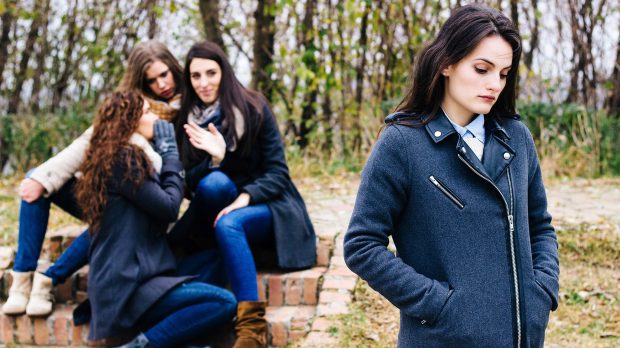 Sad-girl-with-friends-gossiping-in-background-behind-her-back-shutterstock