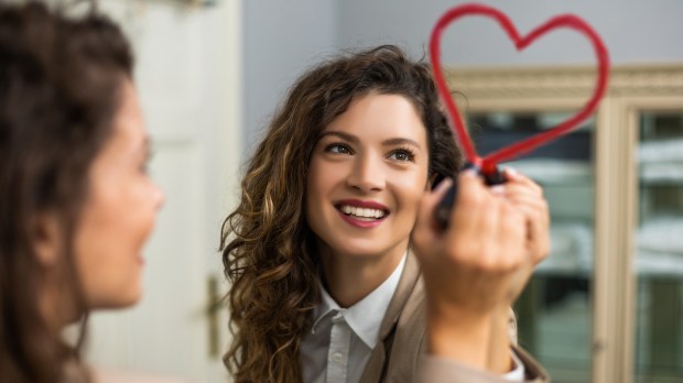 Businesswoman is drawing heart with lipstick on the mirror while preparing for work