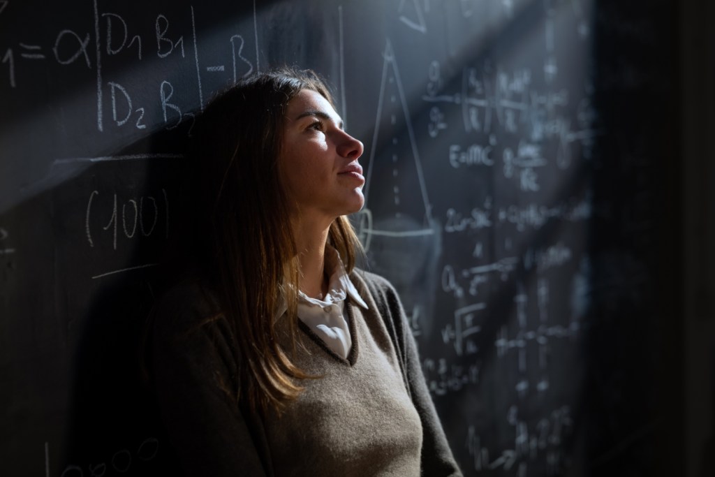 Zamyślona studentka matematyki przed tablicą zapisaną wzorami matematycznymi i fizycznymi